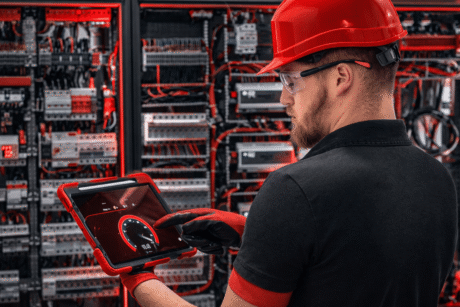 Profissional usando capacete e óculos de proteção analisa um painel elétrico com cabos e equipamentos, utilizando um tablet para monitoramento.