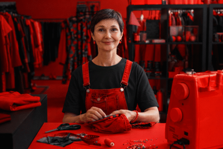 Mulher sentada em uma mesa costurando uma peça vermelha, com máquina de costura, tesoura e materiais de costura ao redor, em um ambiente de ateliê.