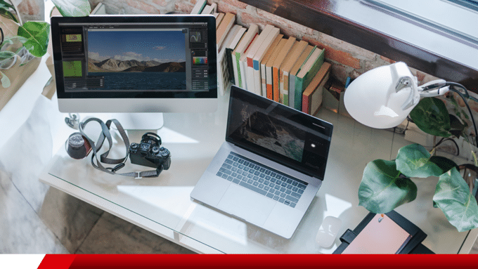Mesa de trabalho com computador, notebook e câmera fotográfica.