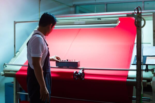 Um profissional opera uma máquina industrial enquanto acompanha o processamento de um tecido vermelho em um ambiente de produção. A cena transmite atenção técnica, controle e atividade fabril.