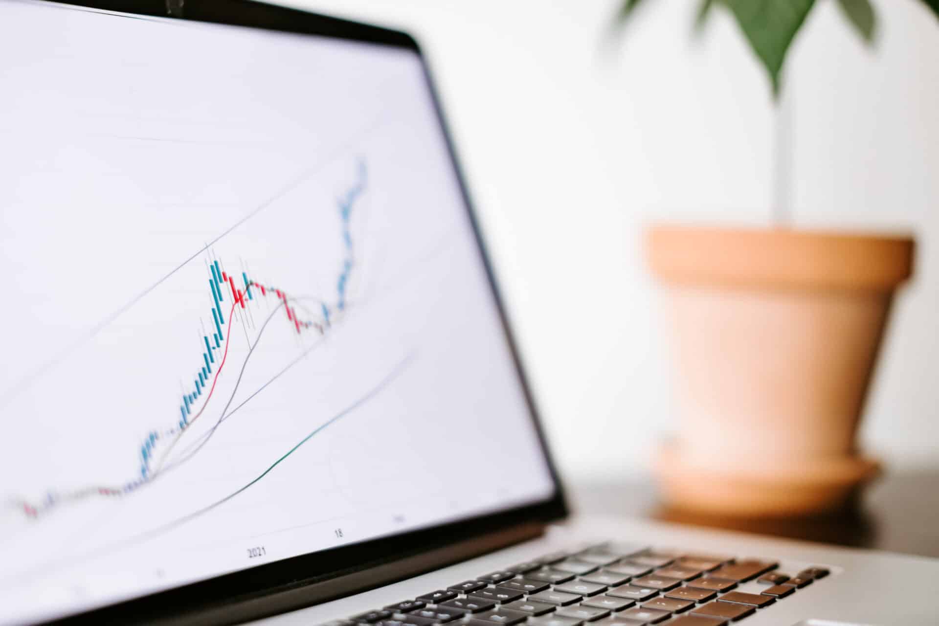 Notebook exibindo um gráfico de mercado financeiro em alta, com um vaso de planta ao fundo.