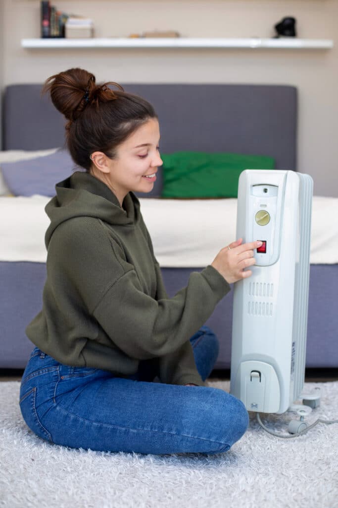 Mulher sentada no chão, em frente a uma cama, acionando o botão de um aquecedor elétrico portátil branco. O aparelho está ligado à tomada e possui um painel com controle de temperatura.