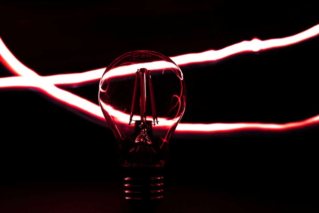 Lâmpada acesa em destaque sobre fundo preto, iluminada por reflexos vermelhos e envolvida por feixes de luz que representam energia elétrica e iluminação.