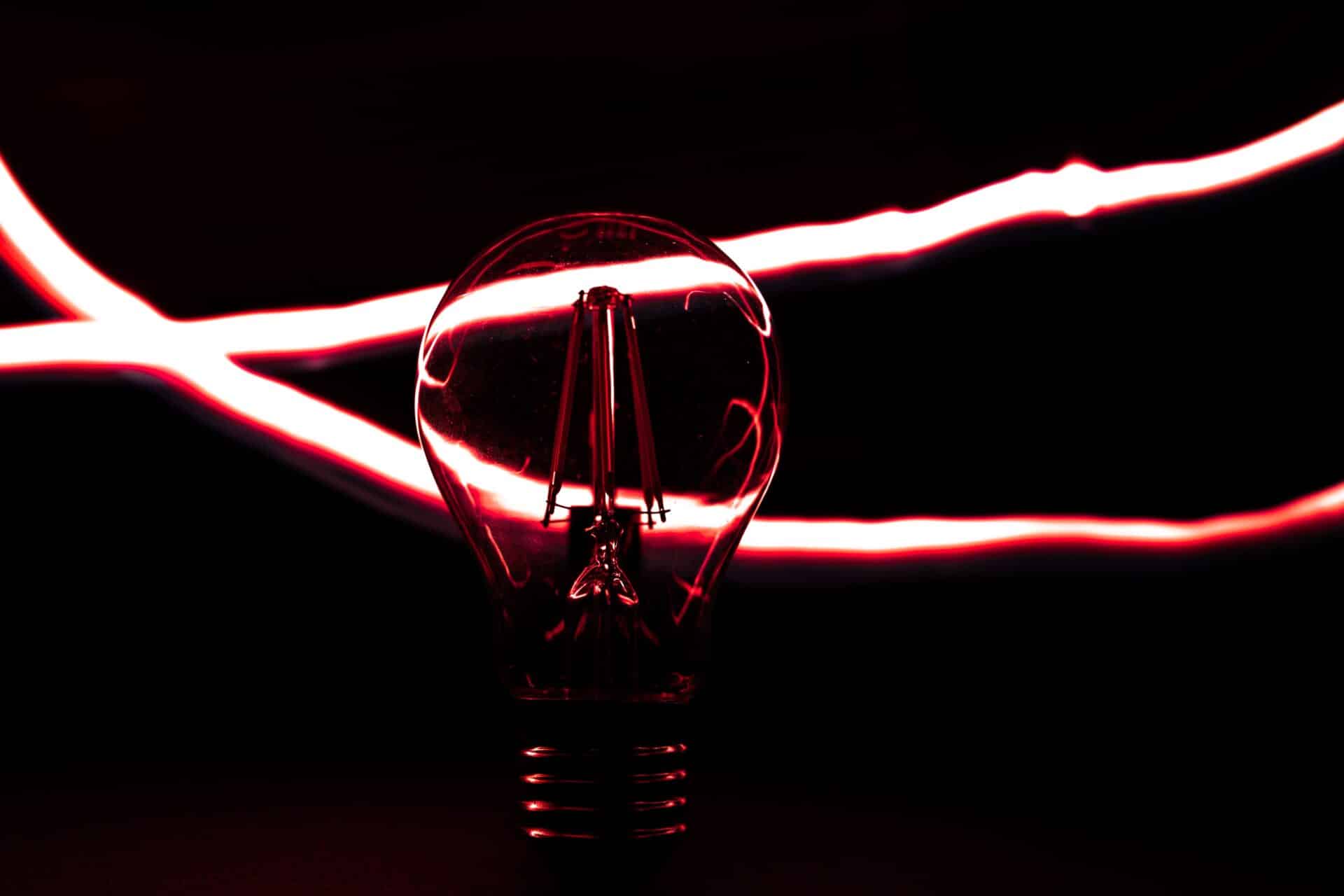 Lâmpada acesa em destaque sobre fundo preto, iluminada por reflexos vermelhos e envolvida por feixes de luz que representam energia elétrica e iluminação.