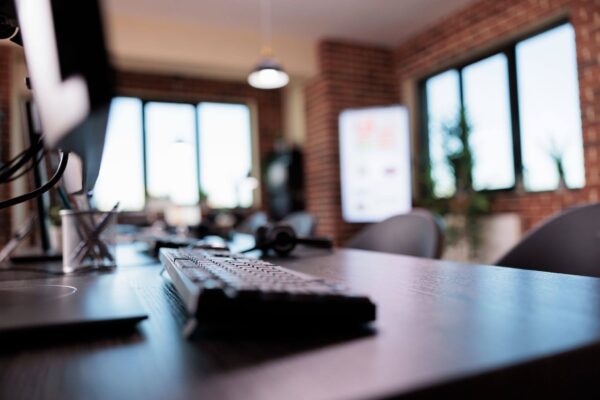 Fotografia de um escritório corporativo moderno, com uma mesa de trabalho em primeiro plano, teclado, notebook parcialmente visível e monitores ao fundo. O ambiente possui paredes de tijolos aparentes, grandes janelas e iluminação natural. A imagem utiliza profundidade de campo, deixando o fundo desfocado e destacando a estação de trabalho.