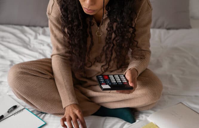 Mulher sentada sobre a cama utilizando um smartphone e organizando documentos, com calculadora aberta na tela e materiais de planejamento ao redor.